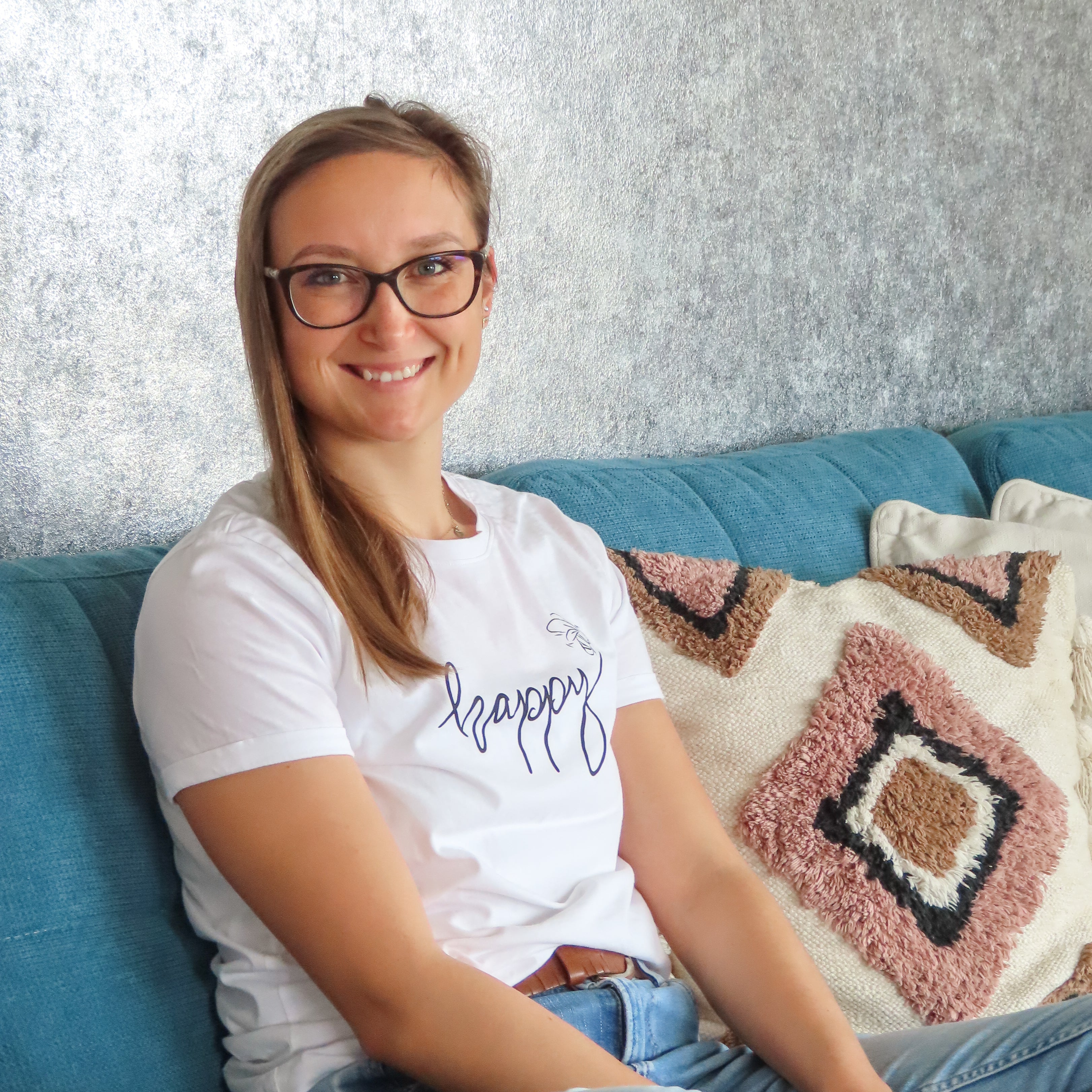 A woman with glasses sitting on a teal couch, wearing a white t-shirt with 'happy' written on it in blue script, paired with light blue jeans.